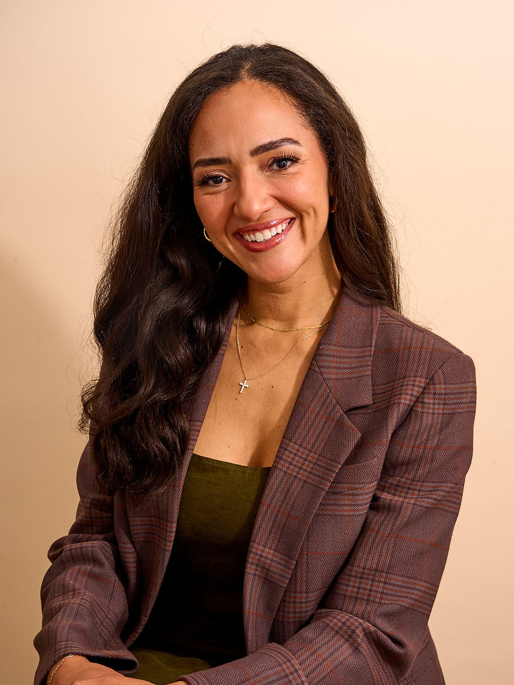 Smiling woman with long dark hair wearing a plaid blazer and green top, against a beige background.