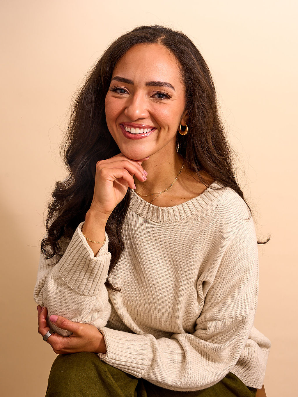 Smiling woman with long dark hair, wearing a cream sweater and gold hoop earrings, posing with hand near chin.