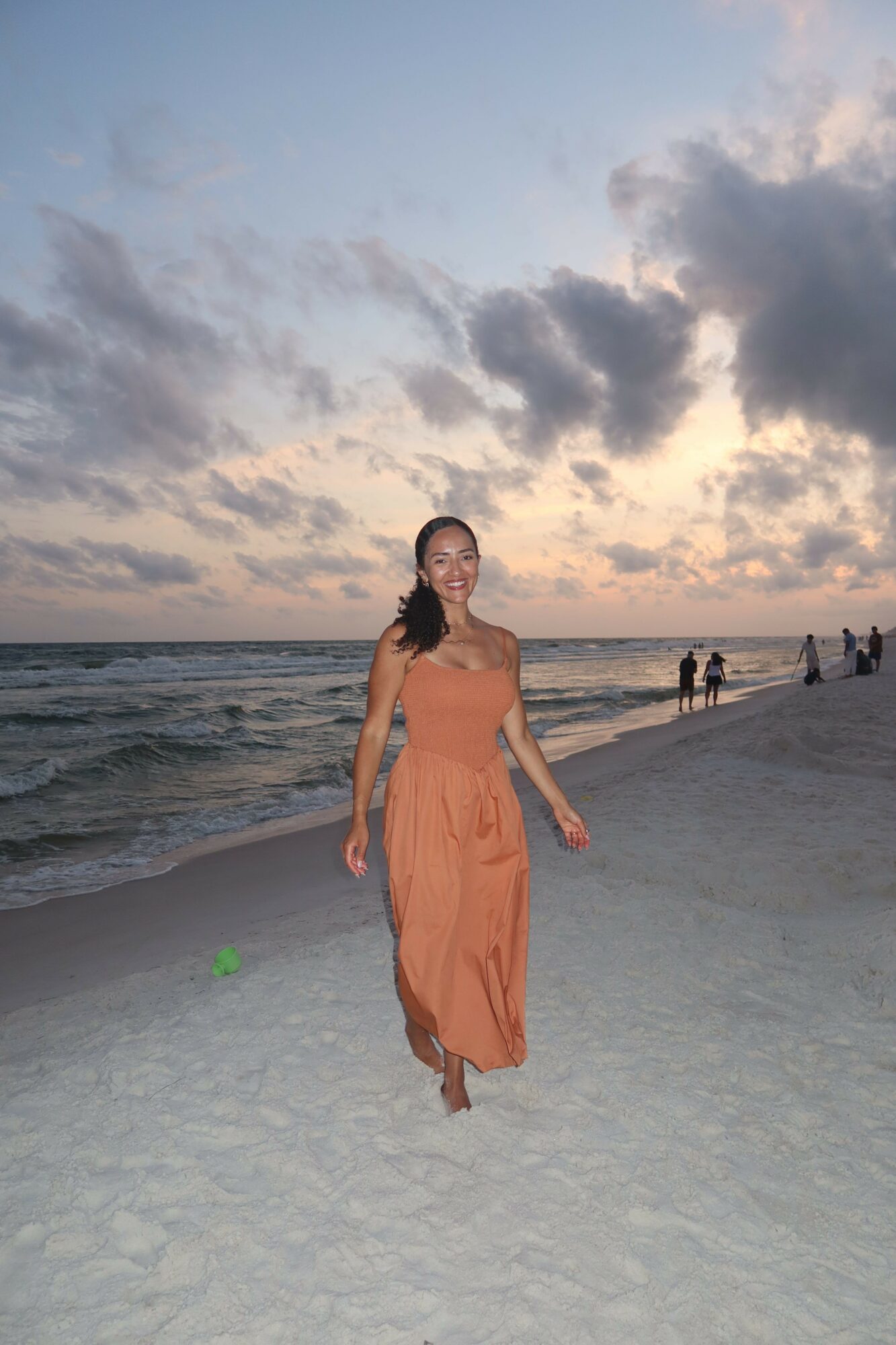 Woman in a long orange dress walking on a beach at sunset, with clouds and water in the background.