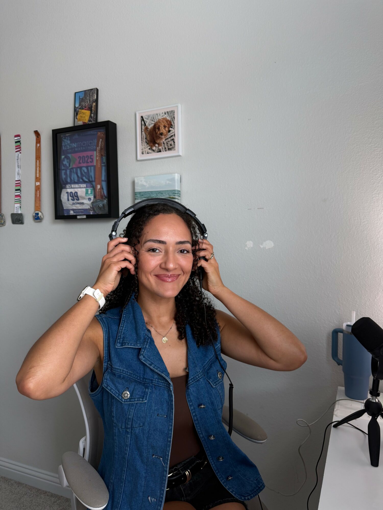 Young woman with headphones smiling, sitting in a room with wall decorations and a microphone, wearing a denim vest.