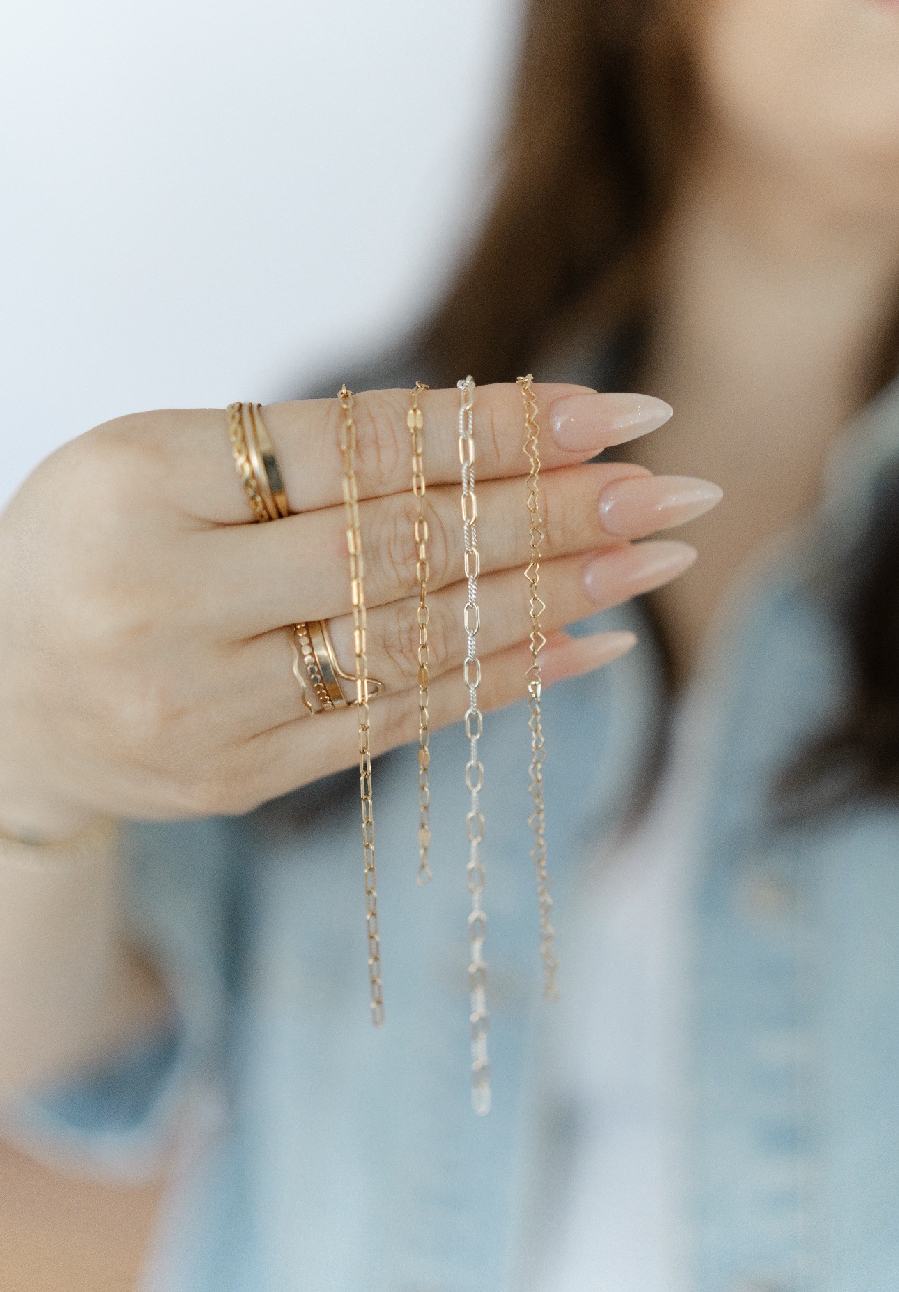 Person's hand with multiple rings and delicate necklaces, blurred background, light blue shirt, long hair, neutral skin tone.