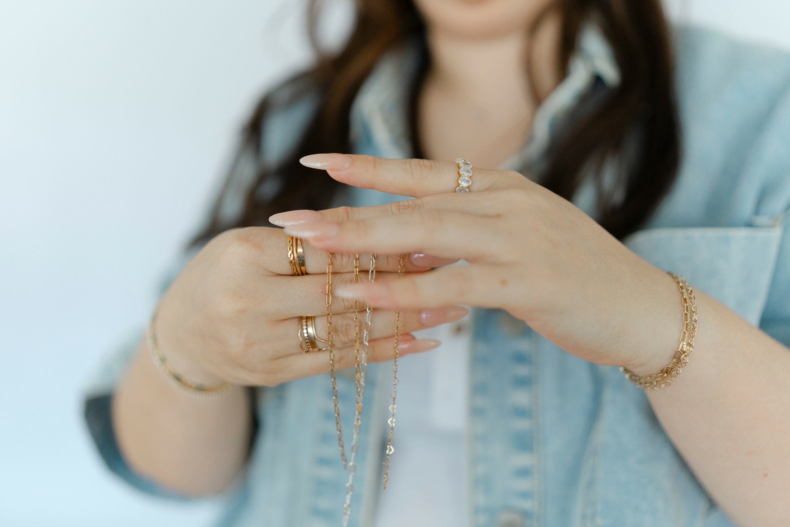 Person's hands with multiple rings and a bracelet, holding jewelry, wearing a denim jacket, with blurred face and background.