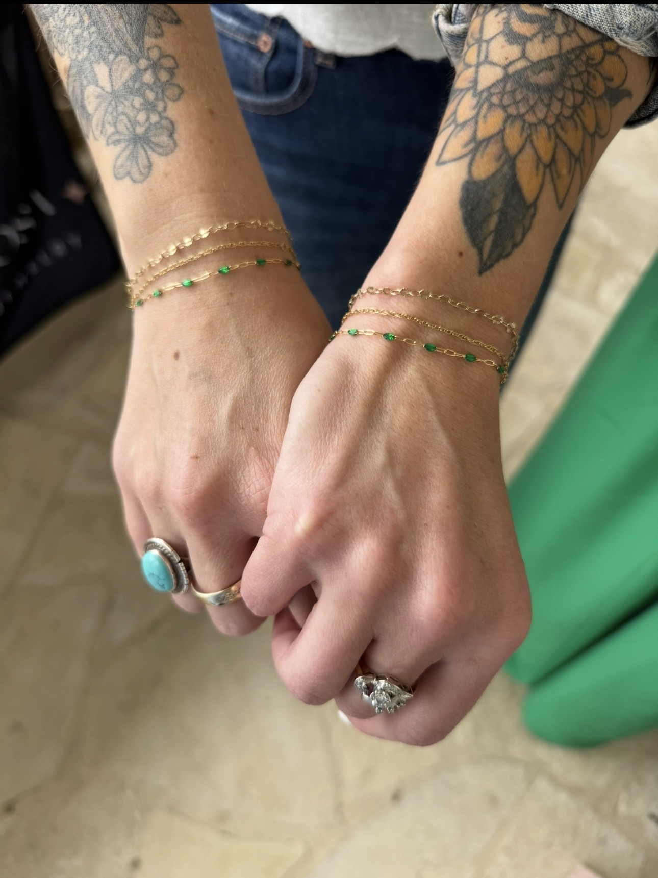 Two hands with tattoos and jewelry, holding each other, showing rings and bracelets, against a blurred background.