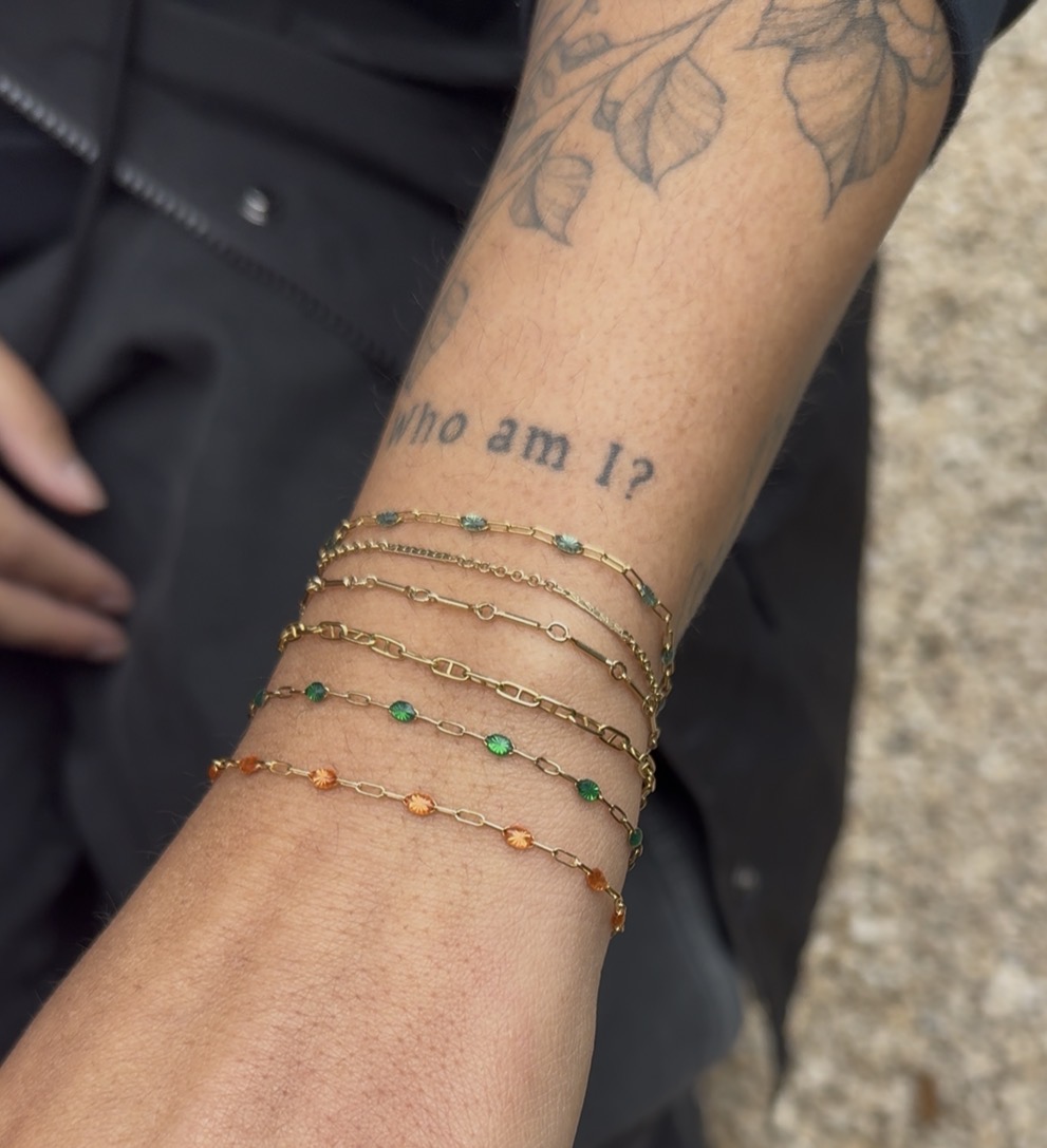 Person's arm with tattoos and four gold bracelets with colorful beads, outdoors on a gravel surface.
