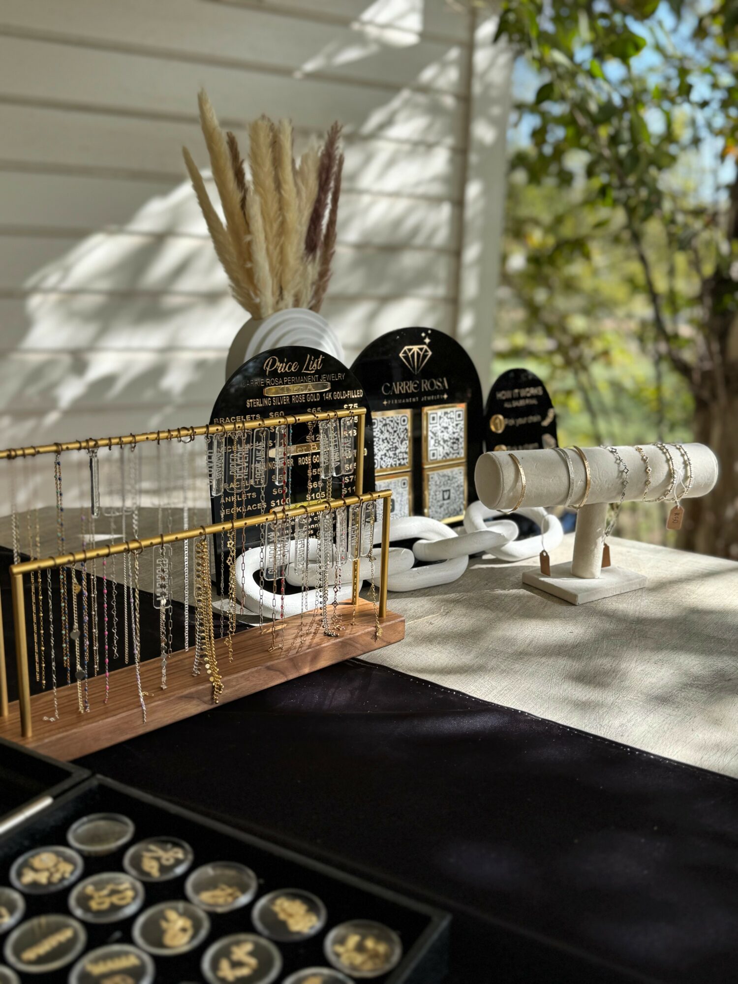 Display of jewelry and decorative items on a table outdoors, with a white wall and green foliage in the background.