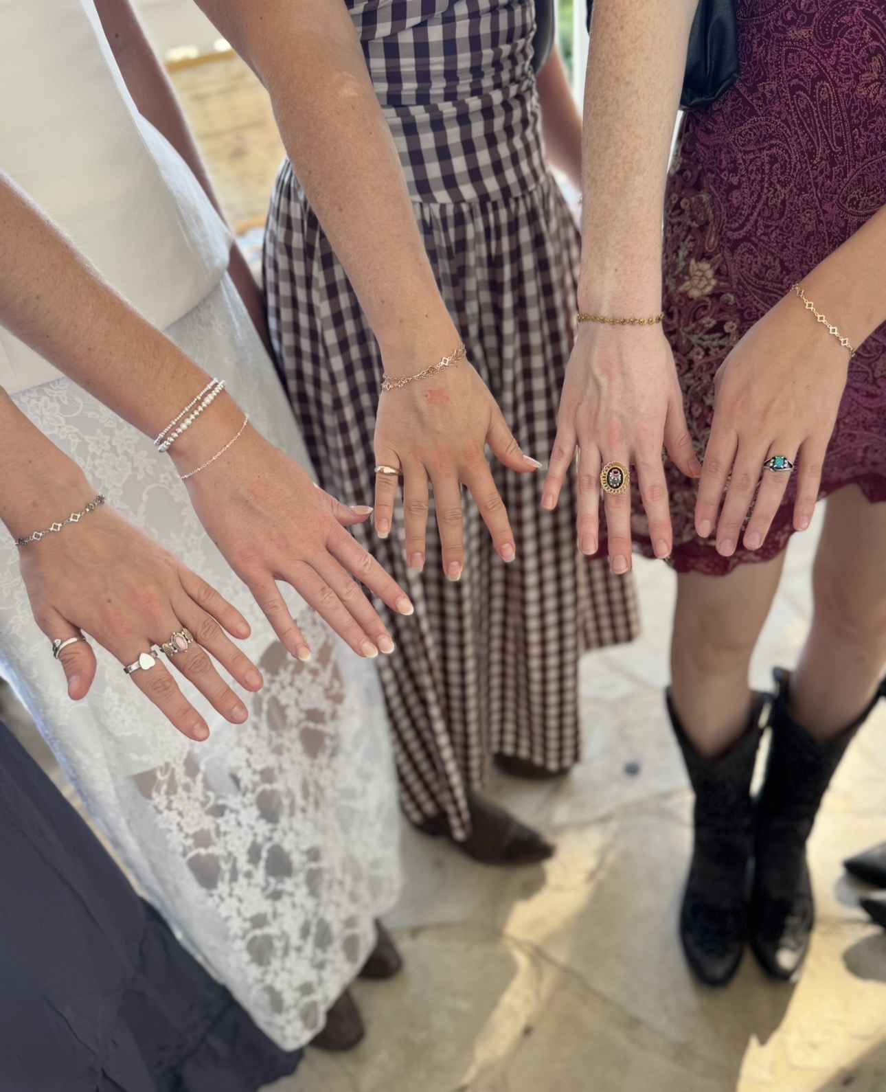 Four women display their hands with jewelry, wearing dresses and boots, standing on a wooden floor.