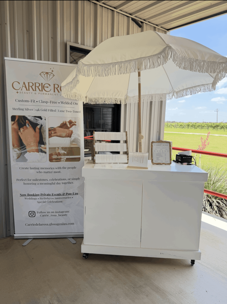 White booth with umbrella and promotional banner, set outdoors with a grassy field and blue sky in background.