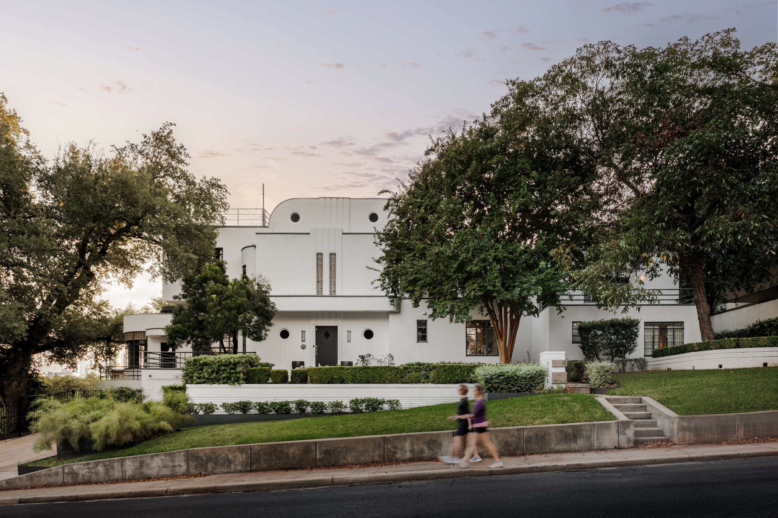 White building with trees and greenery in front, two people walking on sidewalk, sky with clouds, daytime.