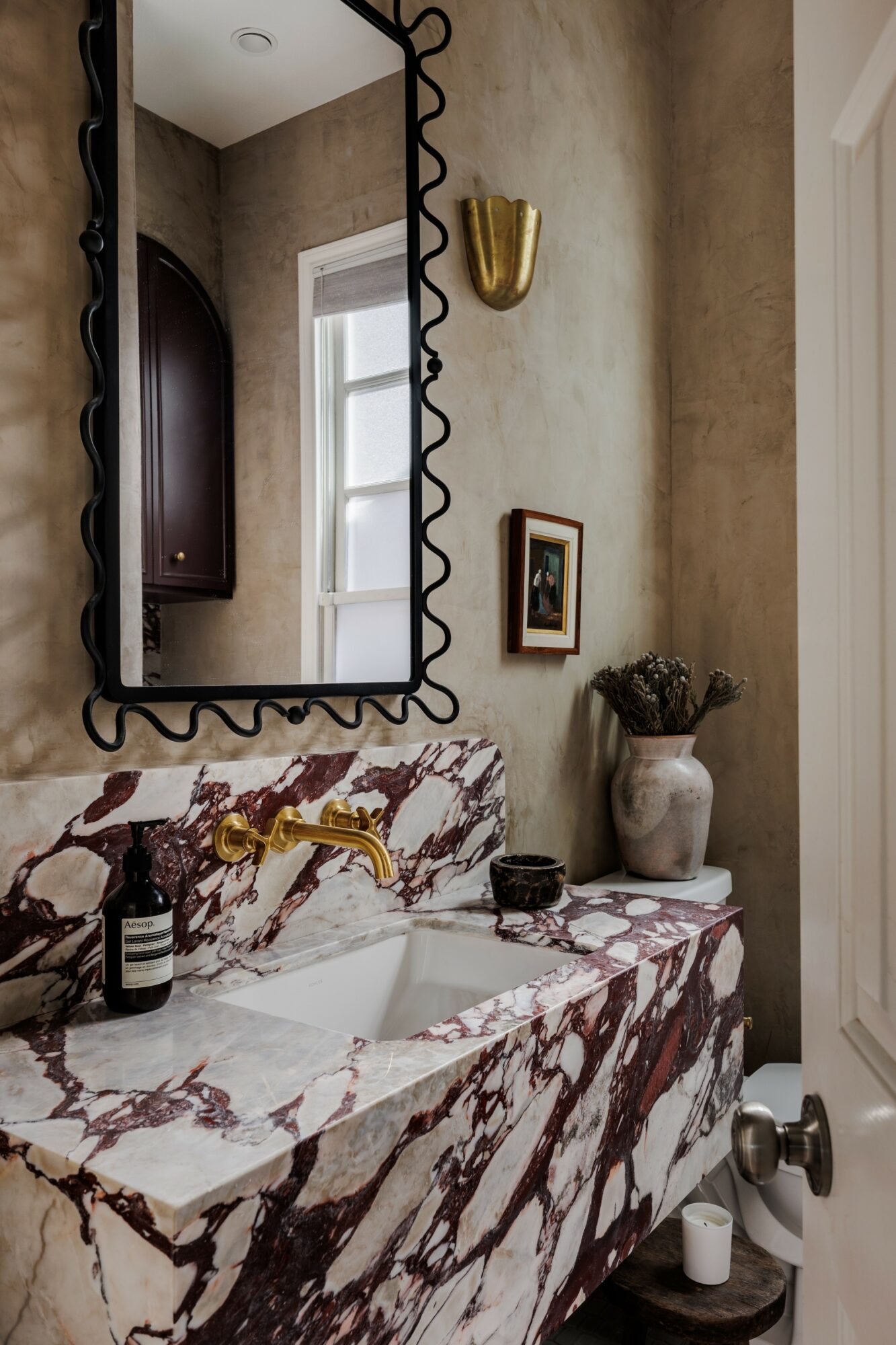Bathroom vanity with marble countertop, mirror, small framed picture, and decorative items, including a vase with dried flowers.