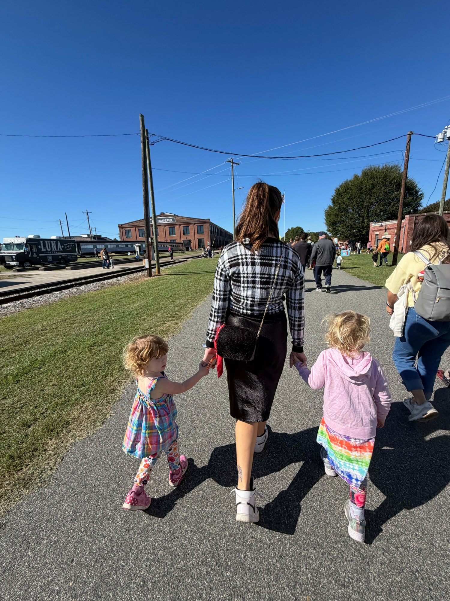 Three children walking on a street with houses and cars, holding hands, under a clear blue sky.