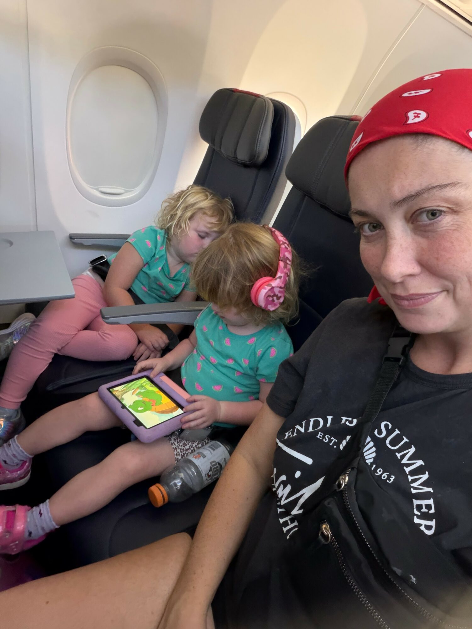 A young woman with a red bandana takes a selfie on an airplane, with two children sitting behind her, one using a tablet.