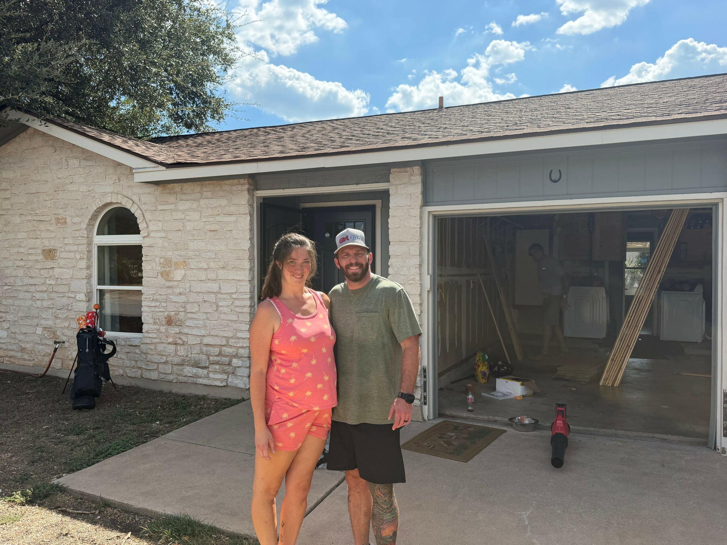 Two people stand outside a garage with a brick house in the background, under a partly cloudy sky.