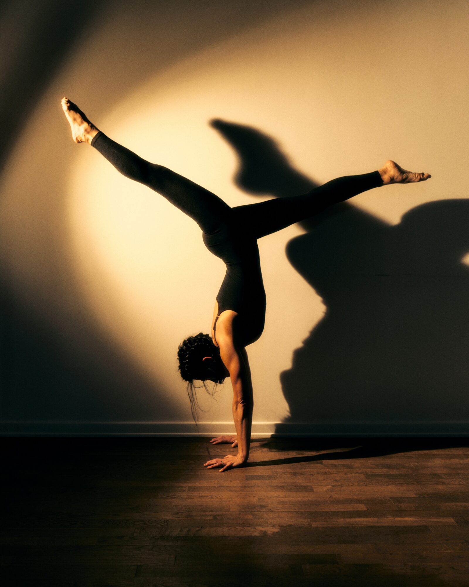 A person balances upside down on one hand with legs extended in a split, casting a shadow on a wall.