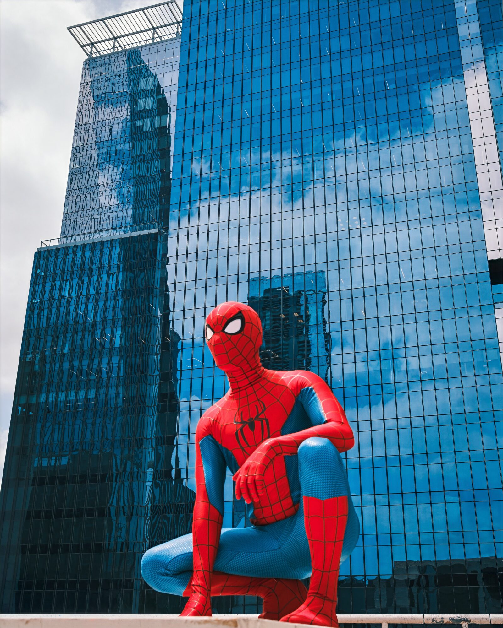 Spider-Man statue crouching in front of a glass building reflecting the sky and clouds.