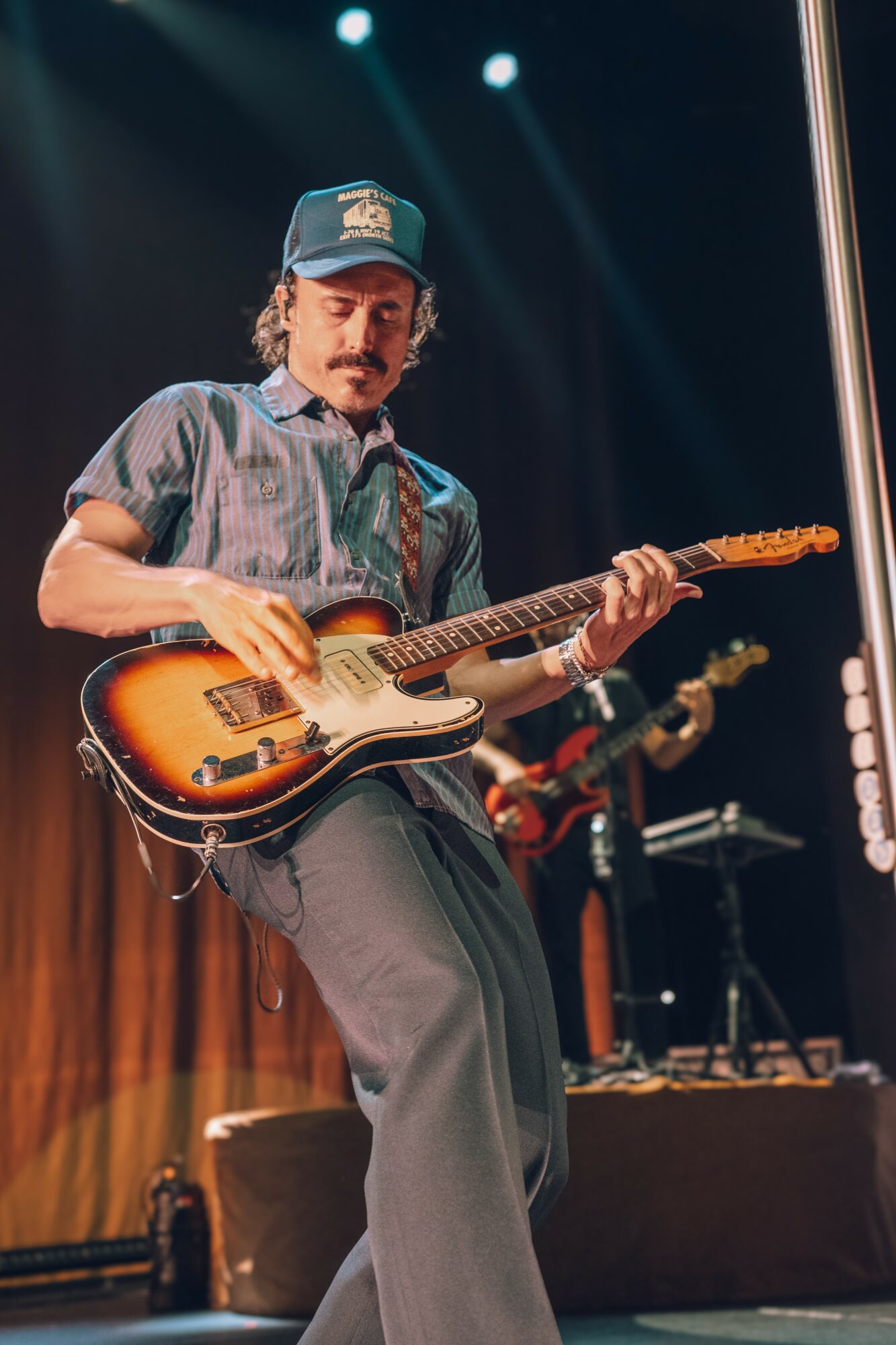 Person playing electric guitar on stage with a hat, wearing a striped shirt and gray pants.