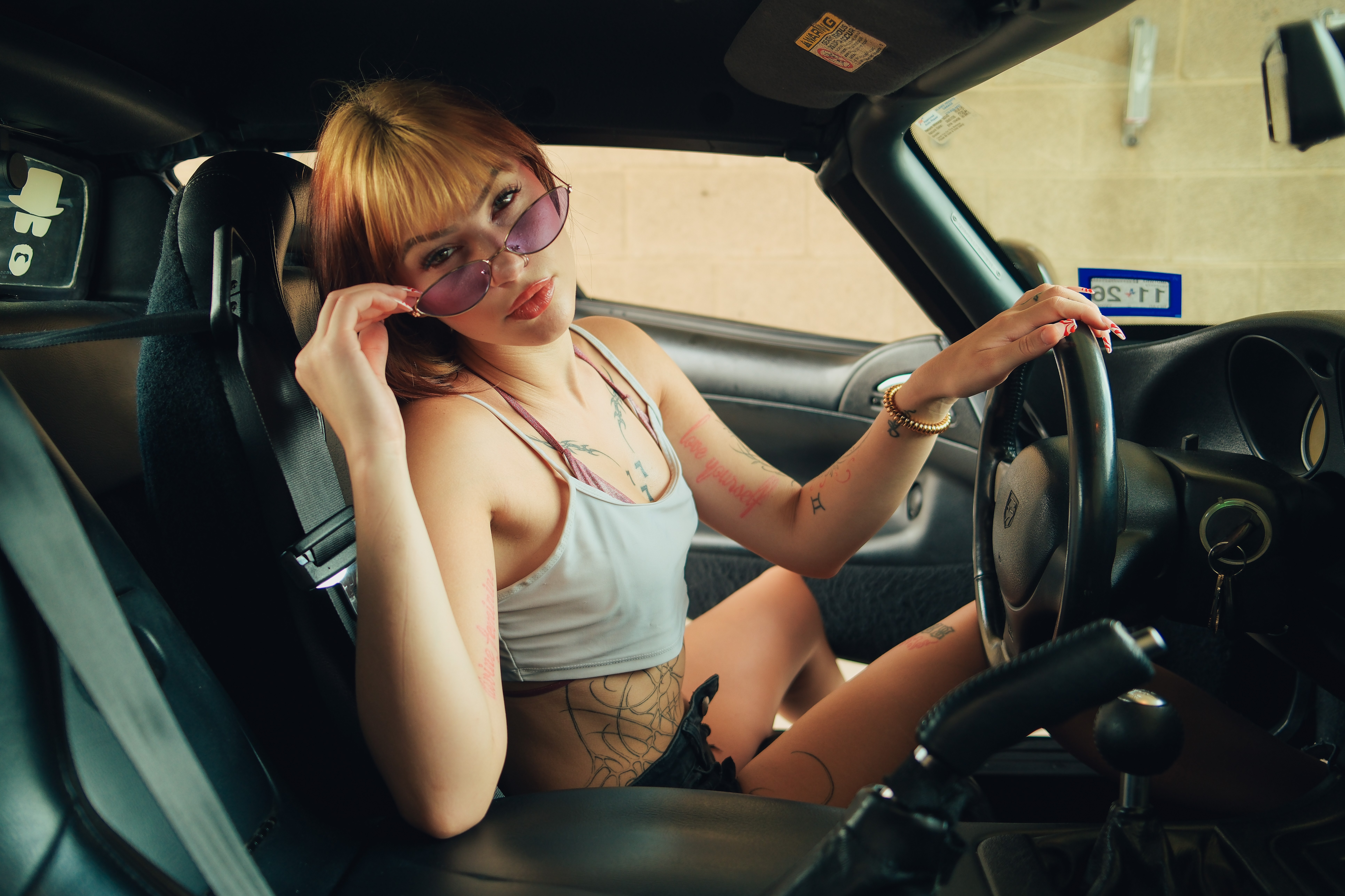 Young woman with glasses sitting in a car, looking at the camera, wearing a white tank top and shorts.