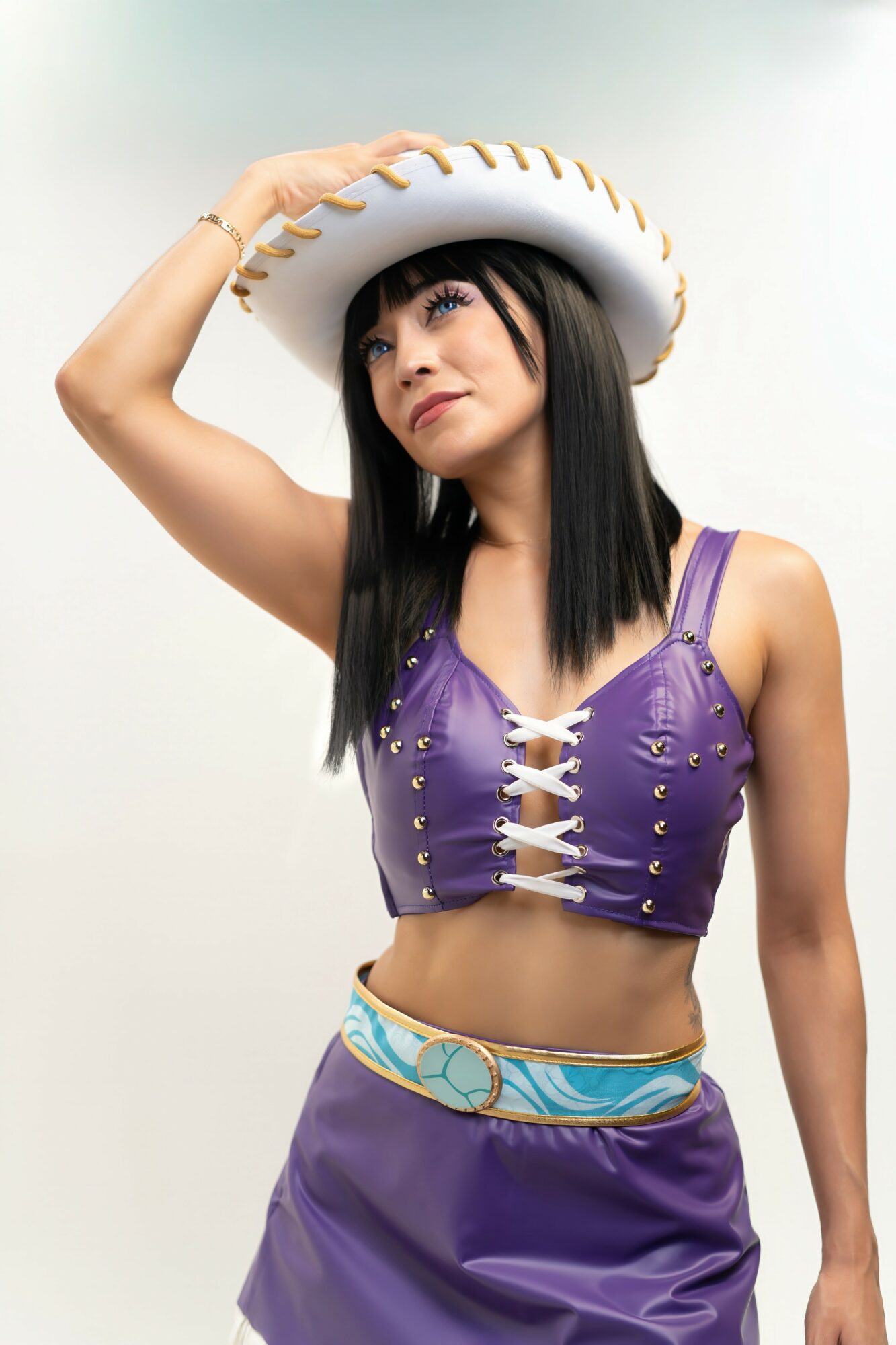 Woman wearing a purple top and a large white hat, holding the hat with her right hand, against a plain background.
