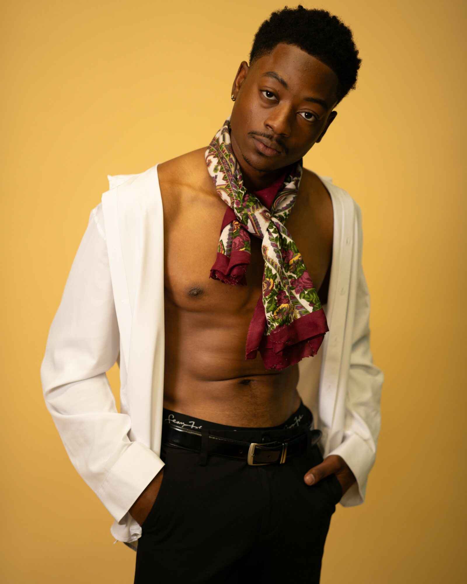 Young man with a scarf around his neck, shirt open, standing against a yellow background.