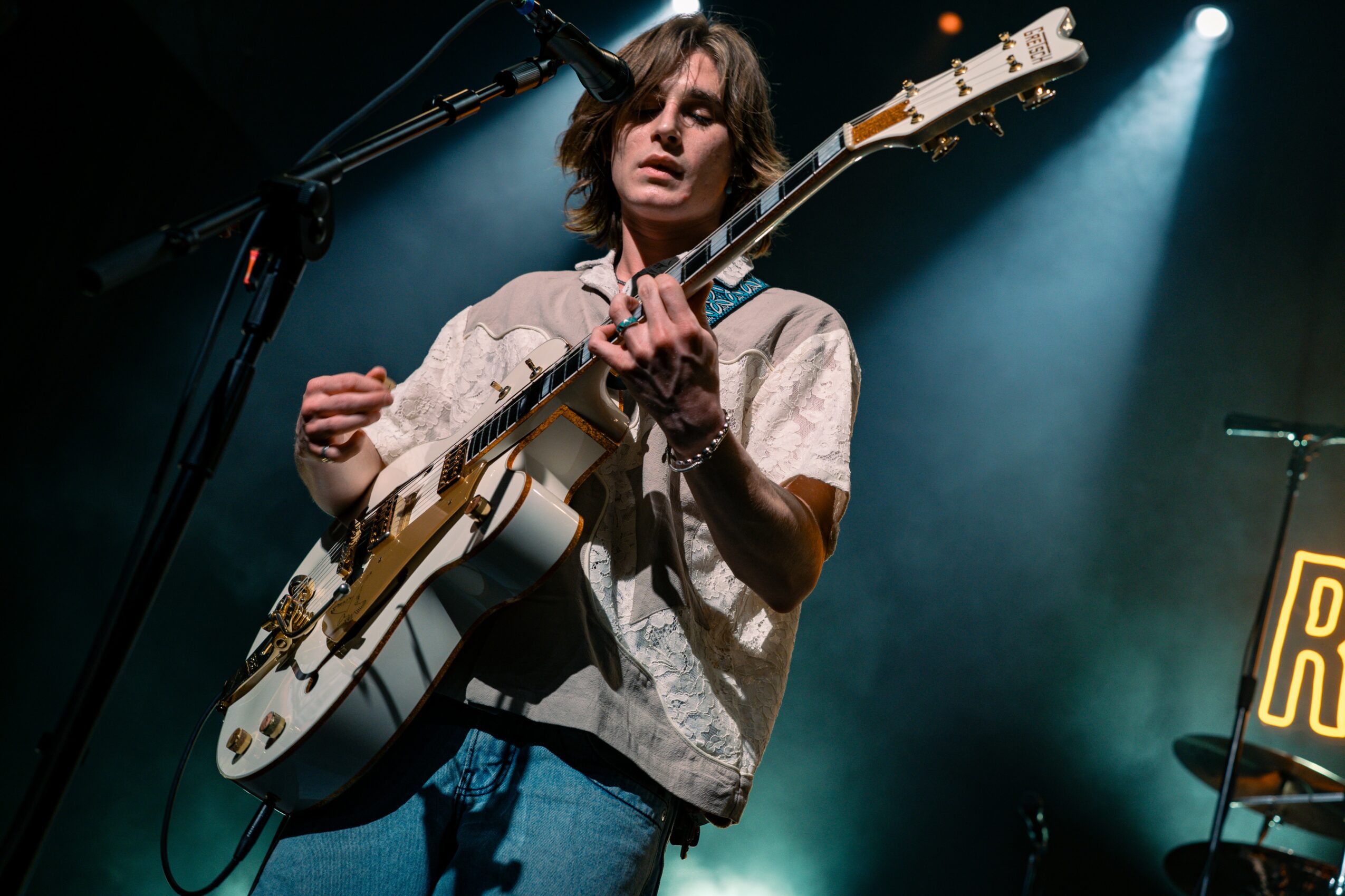 Female musician playing an electric guitar on stage with blue lighting, microphone, and a partially visible drum set in background.