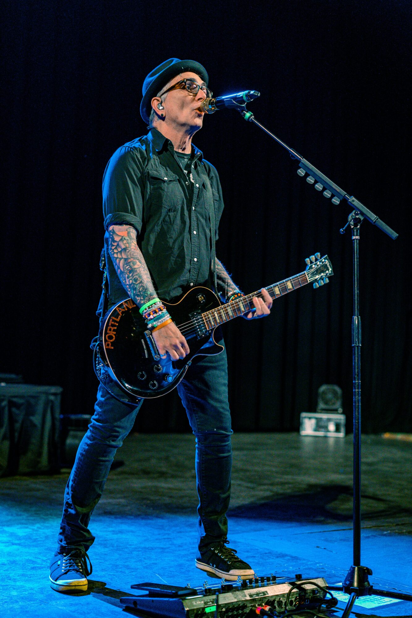 Person with tattoos, glasses, and a beanie playing an electric guitar on stage, singing into a microphone, with dark background.