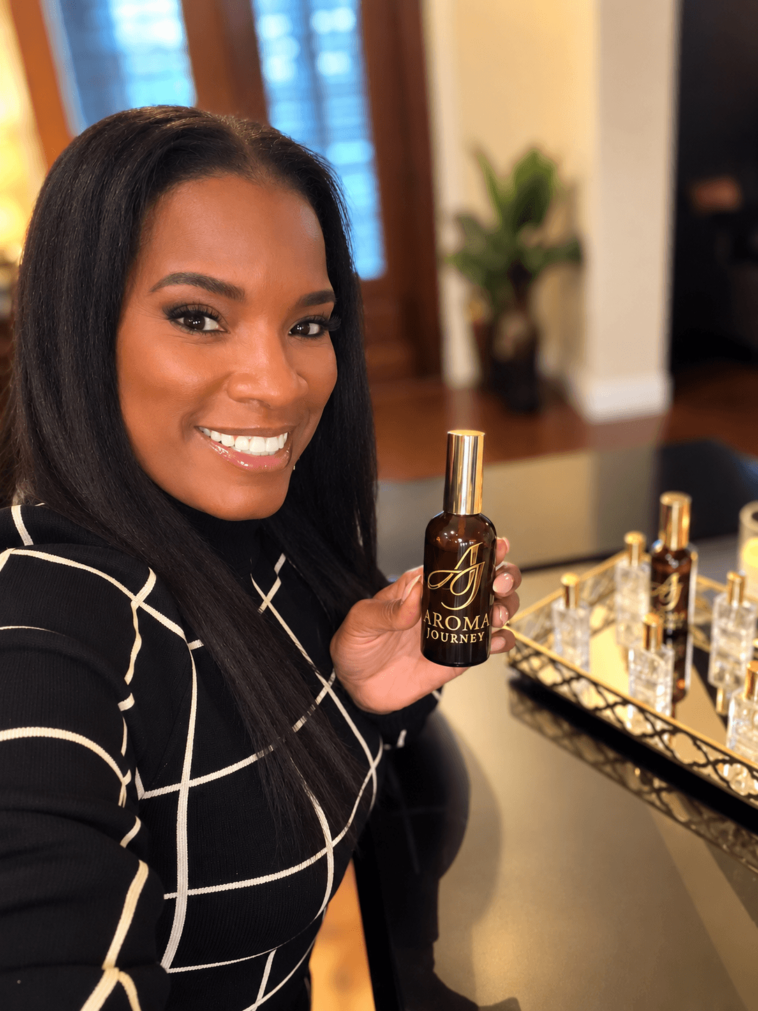 Woman with black hair smiling, holding a small perfume bottle, standing near a tray of perfume bottles, indoors.