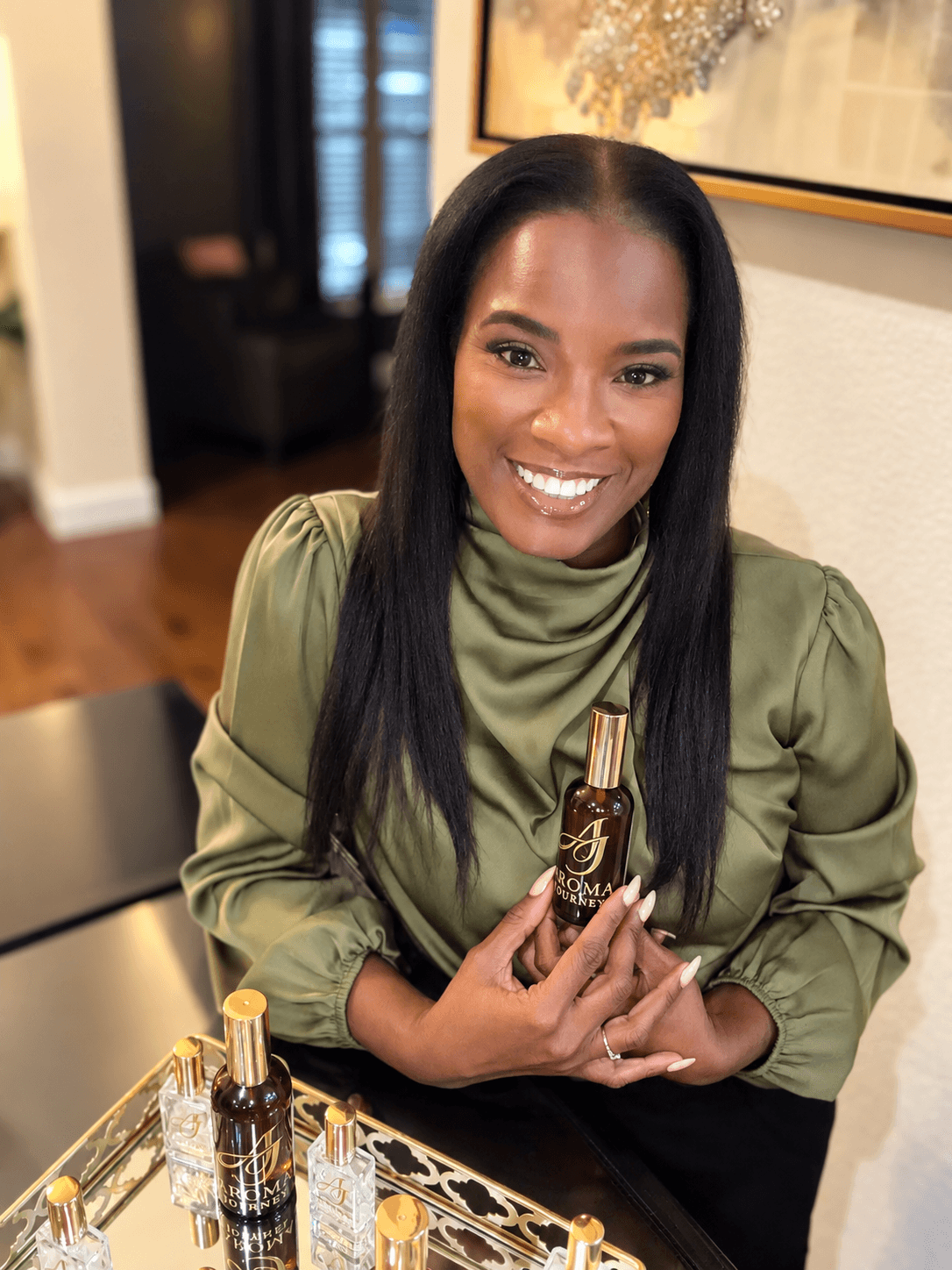 Smiling woman with long black hair holding a small bottle, sitting at a table indoors.