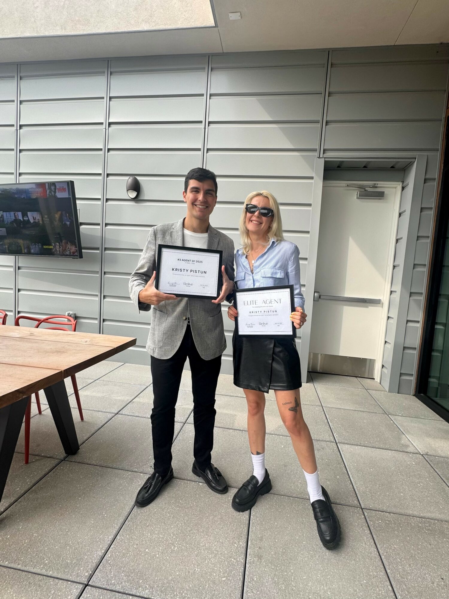 Two people holding certificates, standing indoors in front of a gray wall, smiling, with a table nearby.