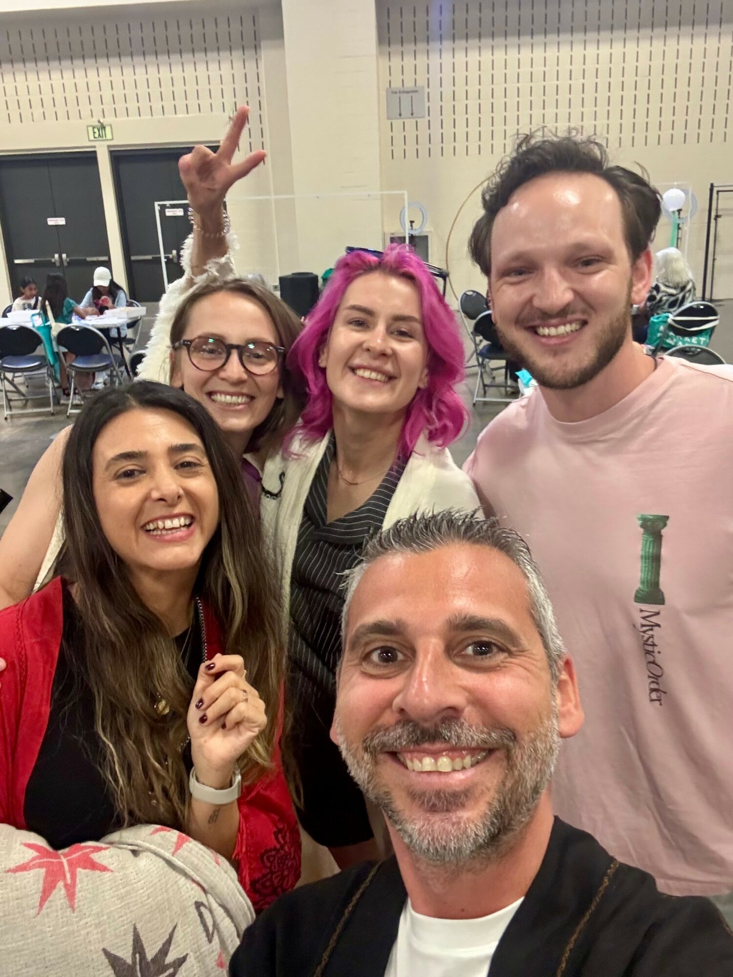 Group of five smiling people taking a selfie indoors, some making peace signs, with tables and chairs in the background.