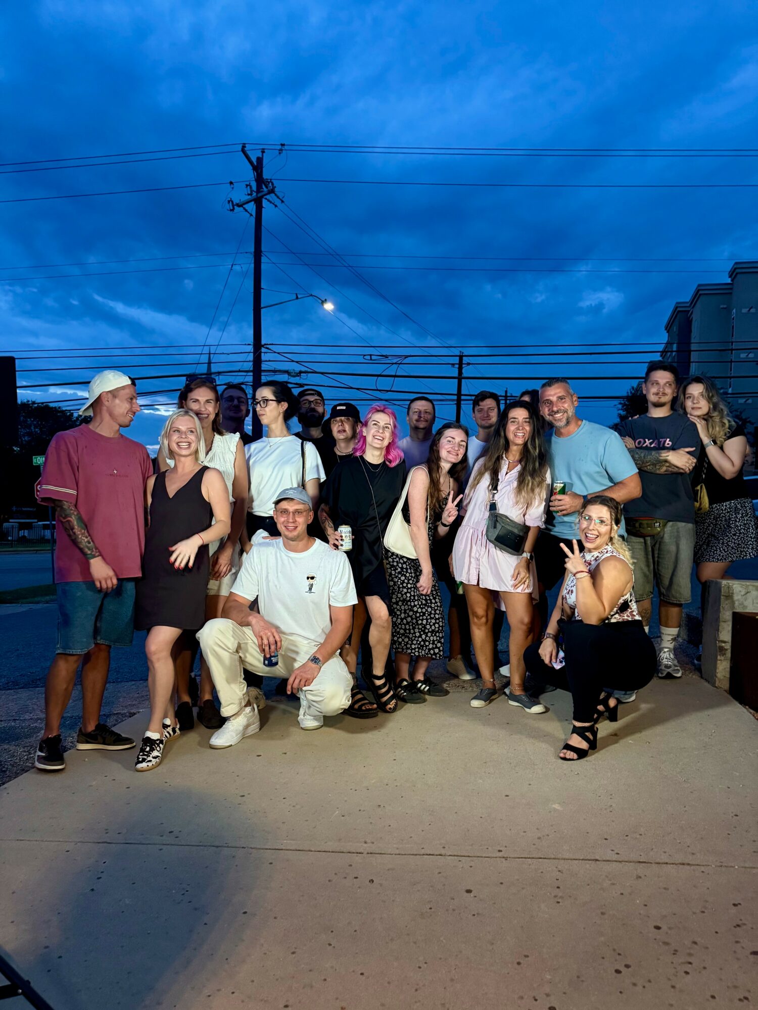 Group of people standing outdoors during evening, some smiling, with a blue sky and power lines in background.
