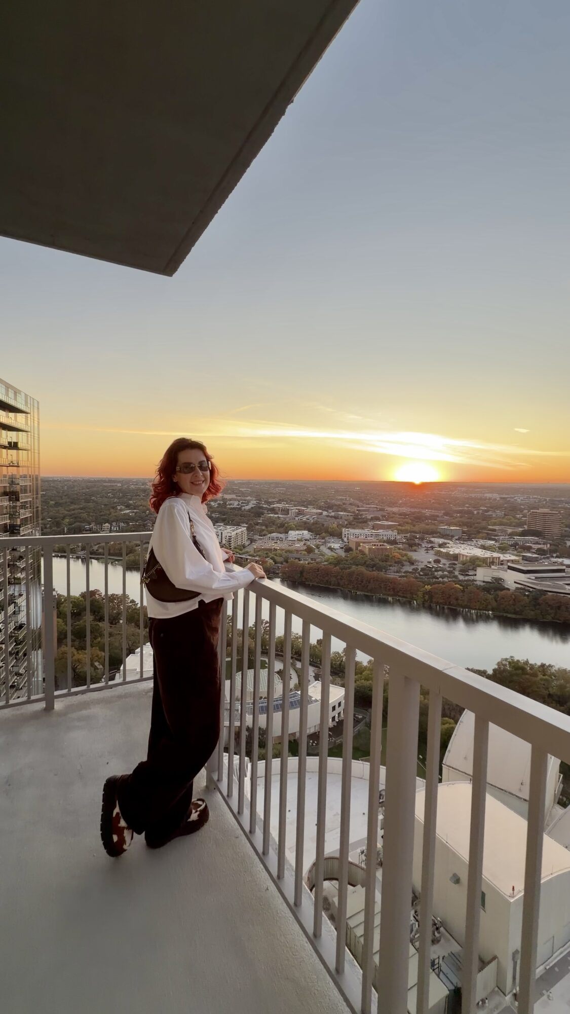 Person with red hair standing on balcony railing overlooking sunset over city and river, wearing white top and dark pants.