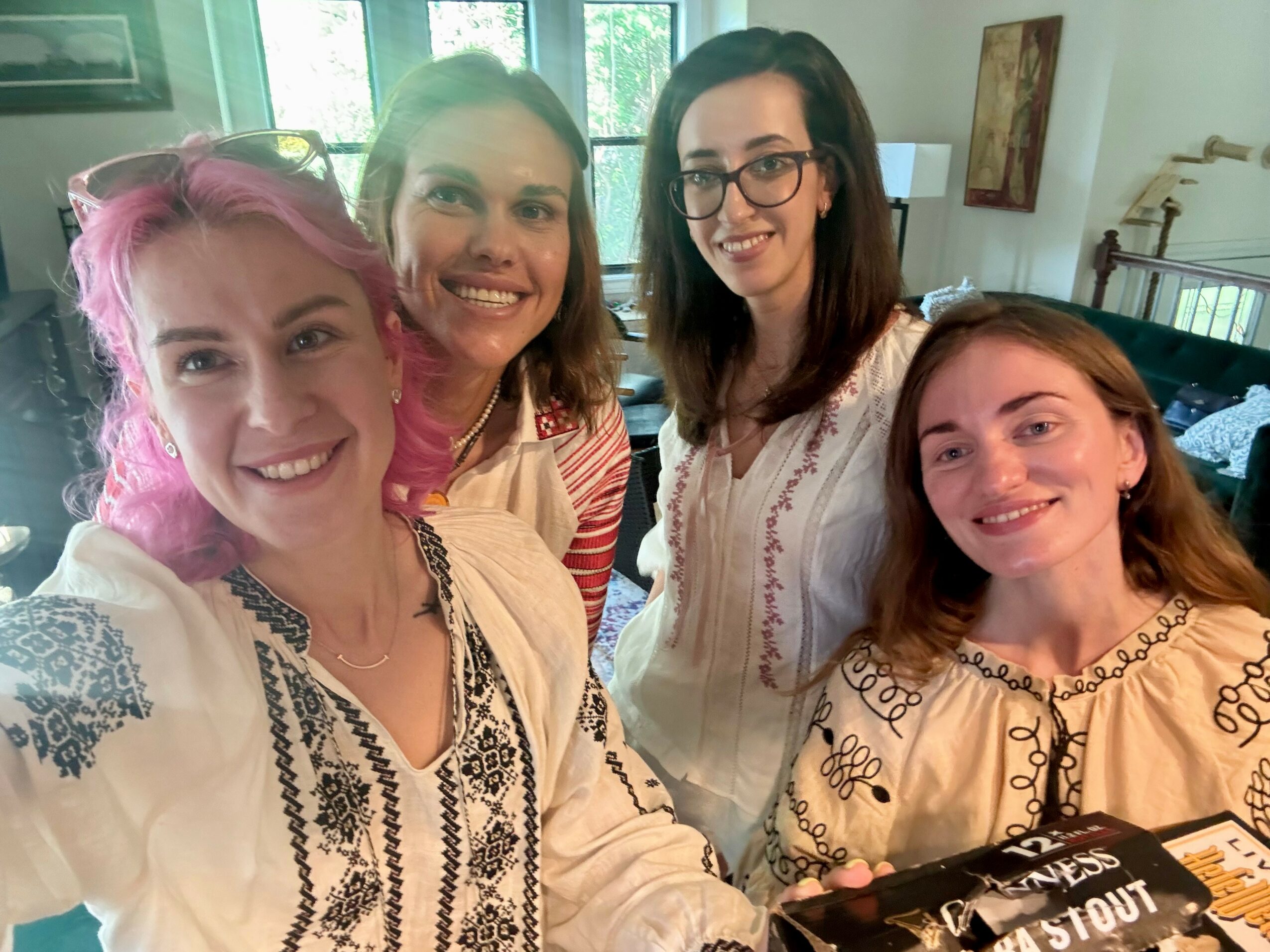 Four women smiling indoors, two with glasses, one with pink hair, in a well-lit room with windows and artwork.