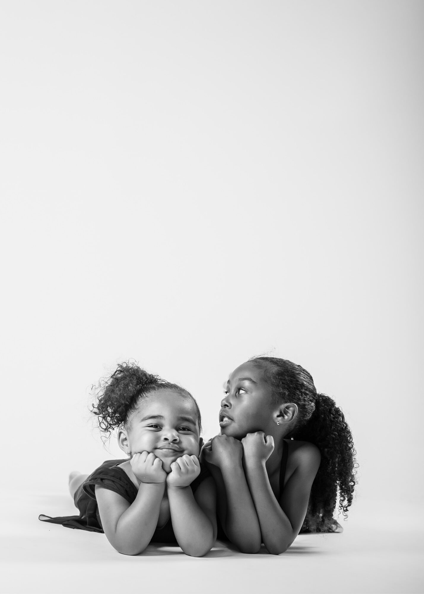 Two young girls lying on their stomachs, facing each other, with their heads resting on their hands, smiling and talking.