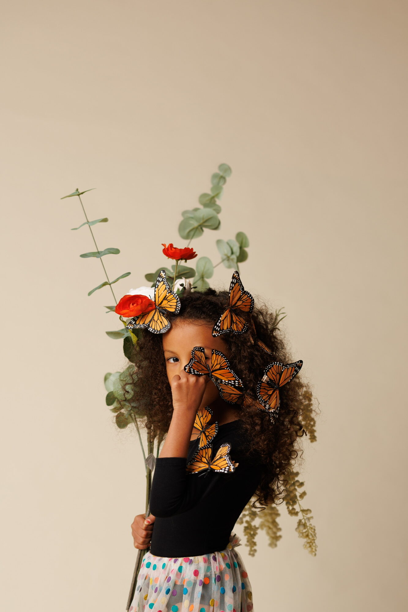 Person with curly hair holding a plant with butterflies and flowers, wearing a black top and colorful skirt.