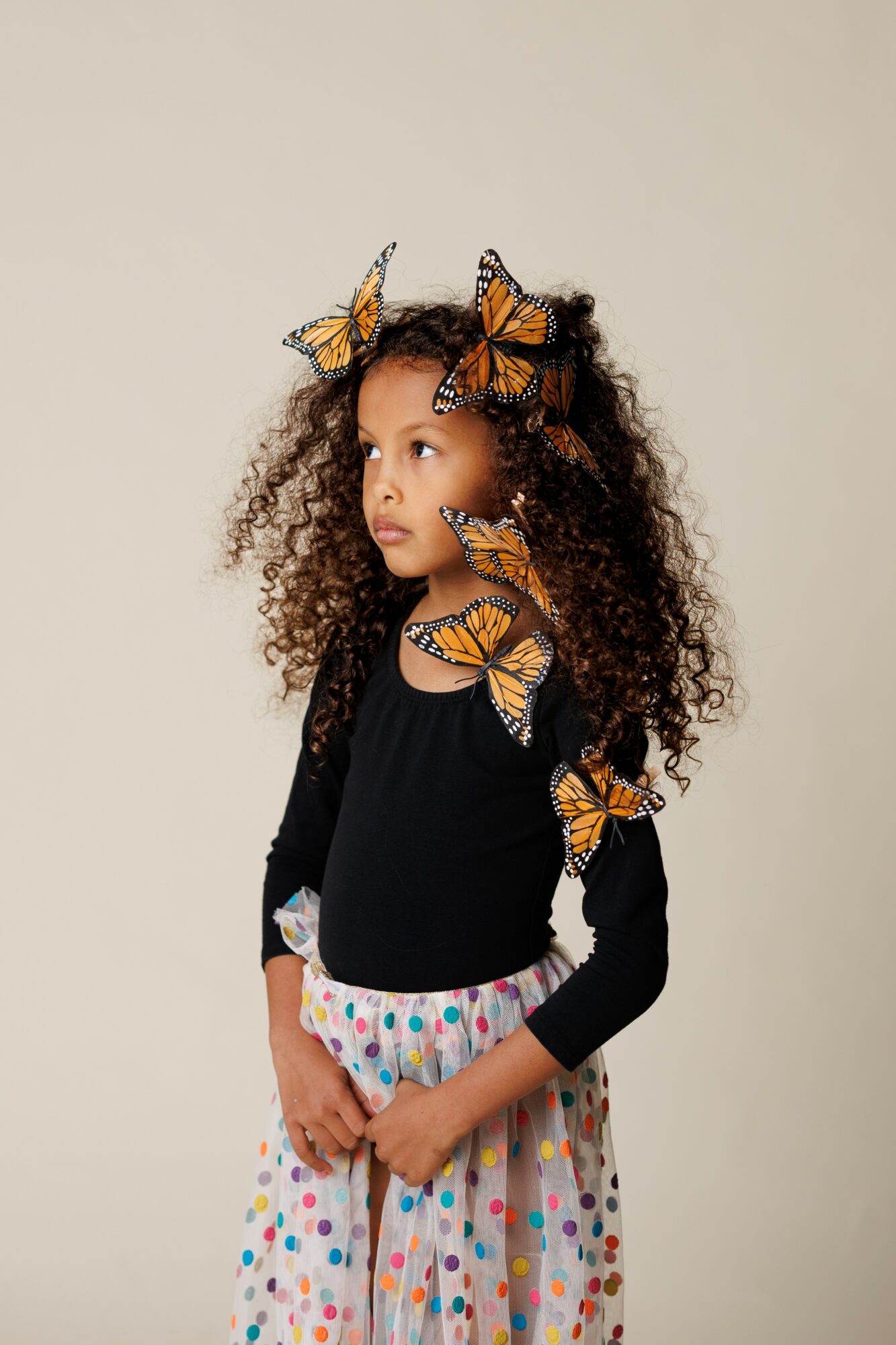 Young girl with curly hair wearing butterfly accessories and a polka dot skirt, standing against a plain background.