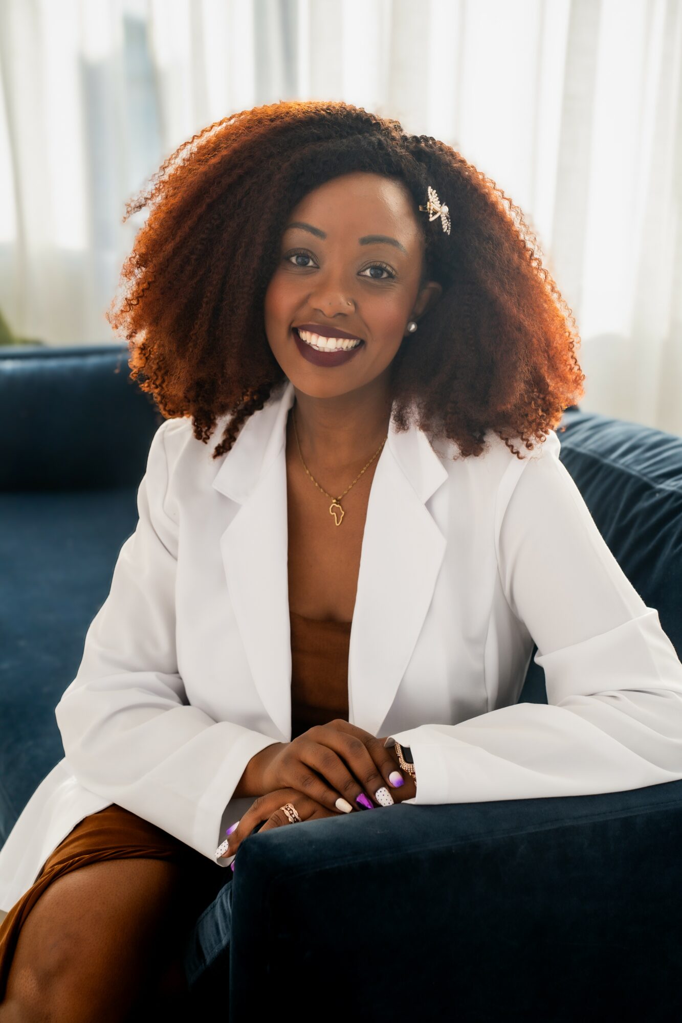 Smiling woman with curly hair wearing a white blazer, sitting on a dark sofa in a bright room.