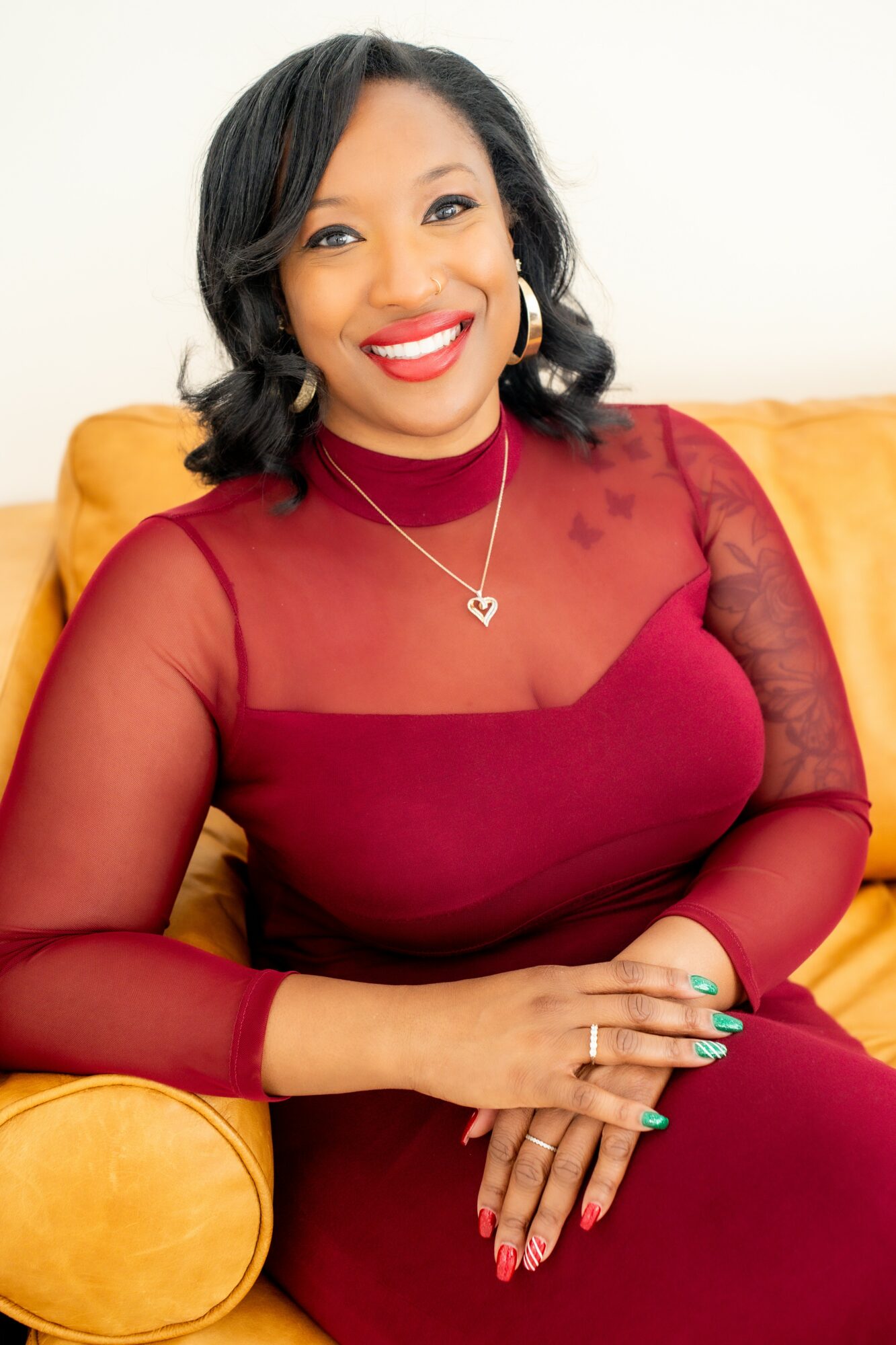 Woman with black hair, red lipstick, wearing a red dress and heart-shaped necklace, sitting on a yellow couch.