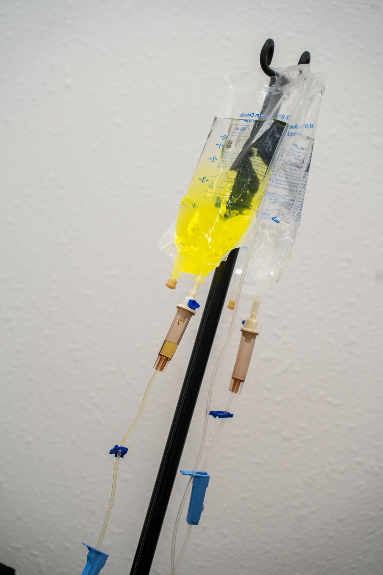 Medical IV drip bag filled with yellow liquid hanging from a stand against a plain wall.
