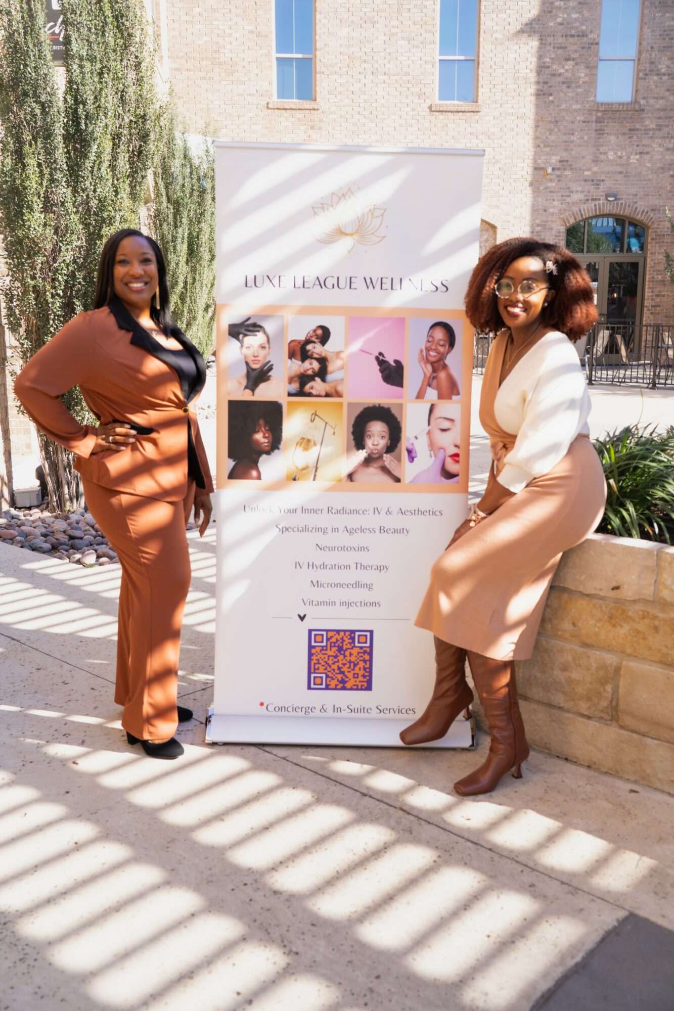 Two women stand beside a promotional poster outdoors, smiling, with a building and trees in the background.