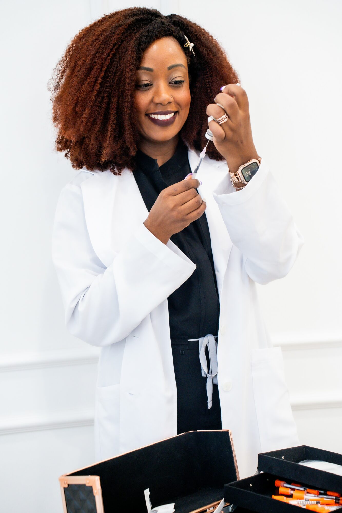 A woman in a white lab coat holding a syringe, smiling, with medical supplies on a table in front.