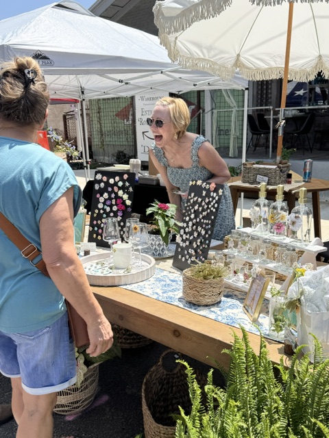 Woman smiling and talking at an outdoor market stall with jewelry and plants, under umbrellas, with another person nearby.
