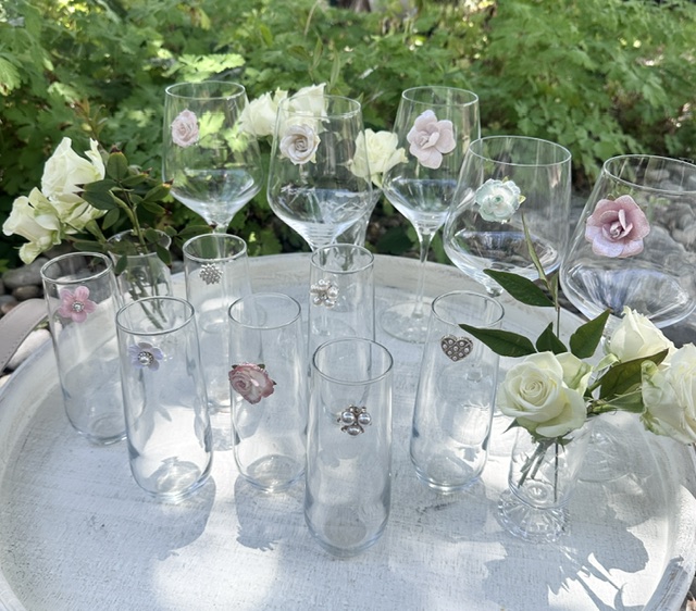 Glassware with flowers on a white table outdoors, greenery in background, some glasses contain flowers, sunlight casting shadows.
