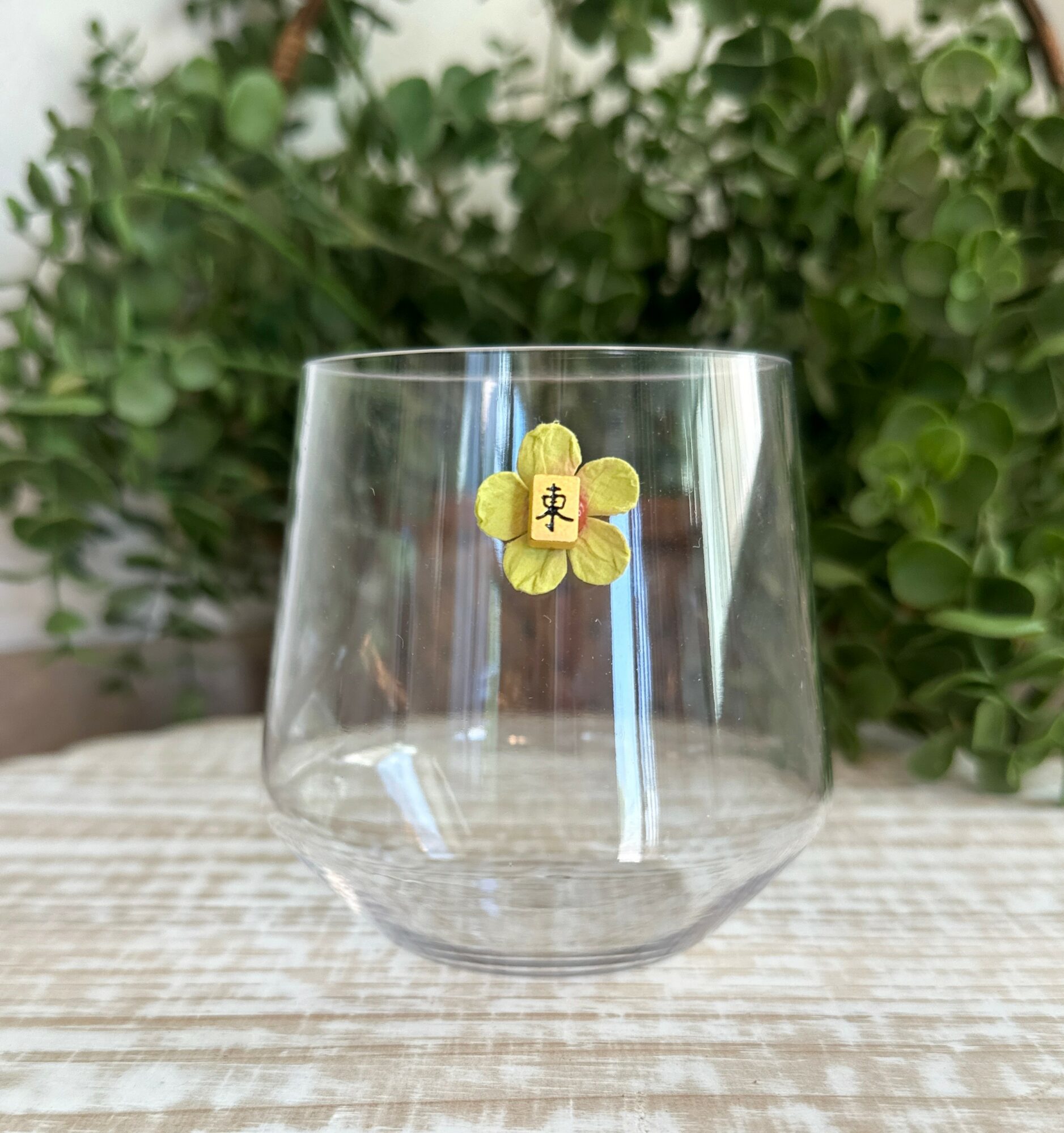 Clear glass with a yellow flower sticker on it, placed on a wooden surface with green foliage background.