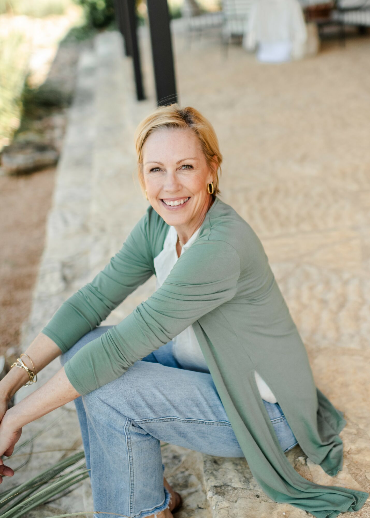 Smiling woman with short blonde hair sitting outdoors on a stone surface, wearing a green cardigan and jeans.