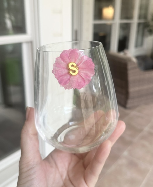 Clear glass held by a hand with a pink flower sticker and a yellow letter S, outdoors with windows and furniture in background.