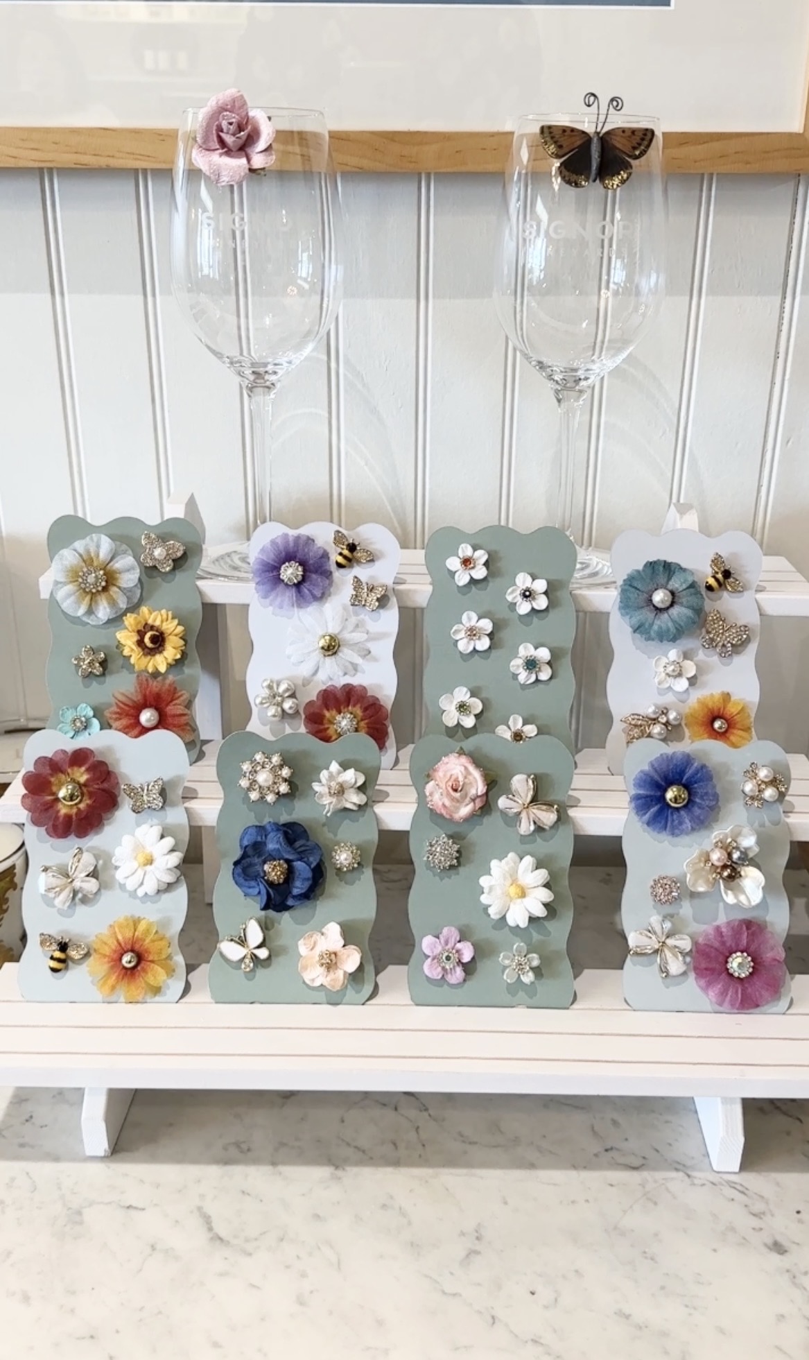 Display of decorated hair clips with flowers and butterflies on a white shelf, with a wooden top and vertical panel background.