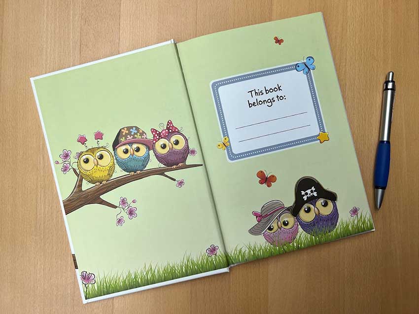 Open children's book showing colorful owls on a branch and a blank page for writing, on a wooden surface with a pen nearby.