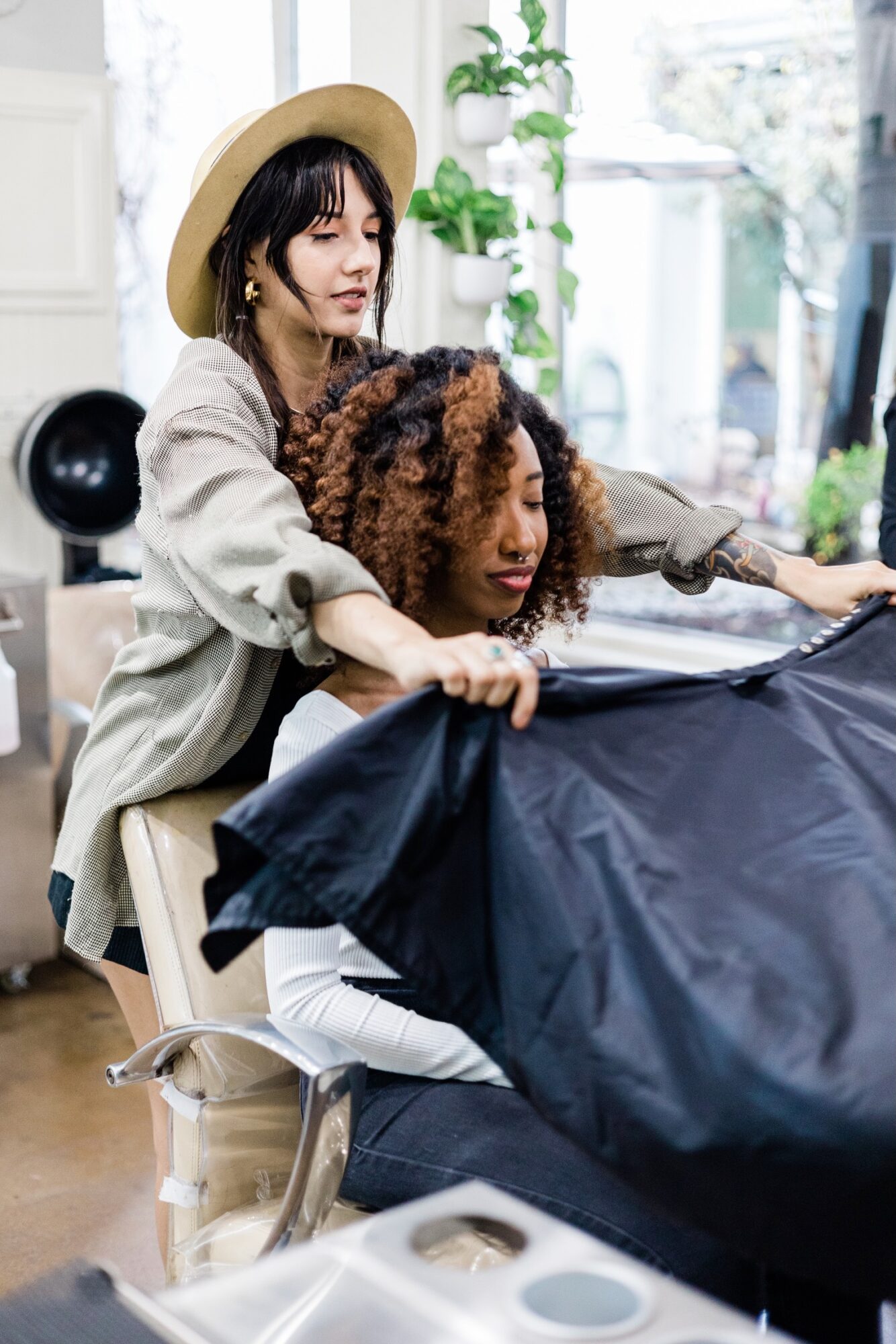 Two women working together at a salon, one with curly hair and the other with straight hair, near a large window.