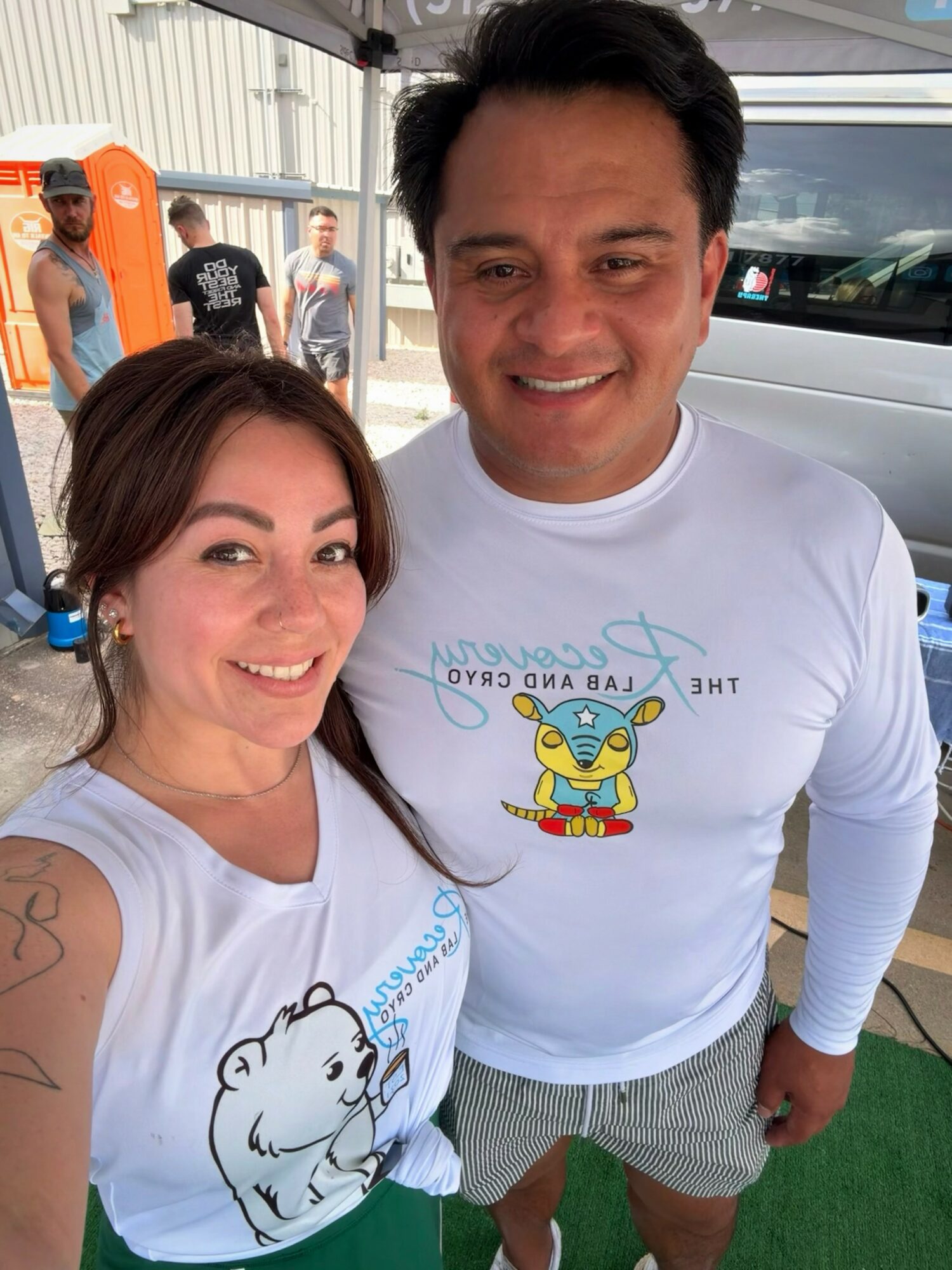 Two smiling people wearing white shirts with cartoon characters, standing outdoors near a white vehicle and a building.
