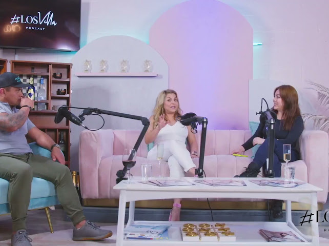 Two women and a man sit on a pink sofa and a blue chair in a podcast studio with microphones, books, and a white table.