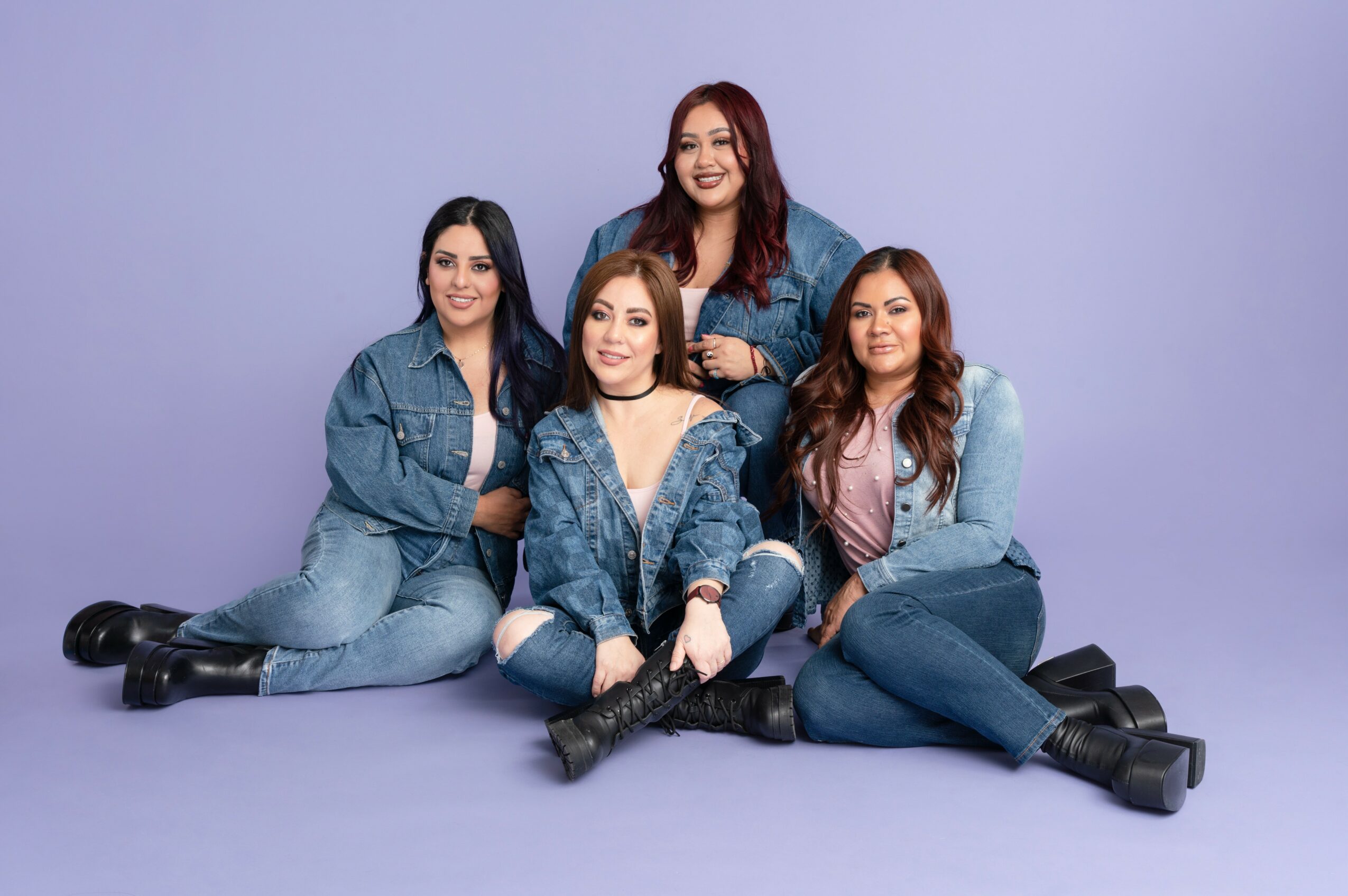 Four women sitting and kneeling on a purple background, smiling, wearing casual denim outfits.